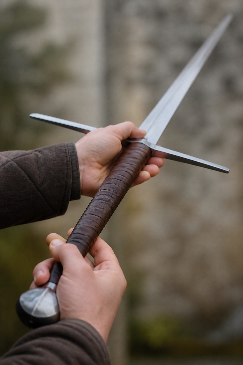 A hand and a half sword held in two hands at chest height, the long leather wrapped grip and the straight cross guard filling the frame, the blade angled away from the camera